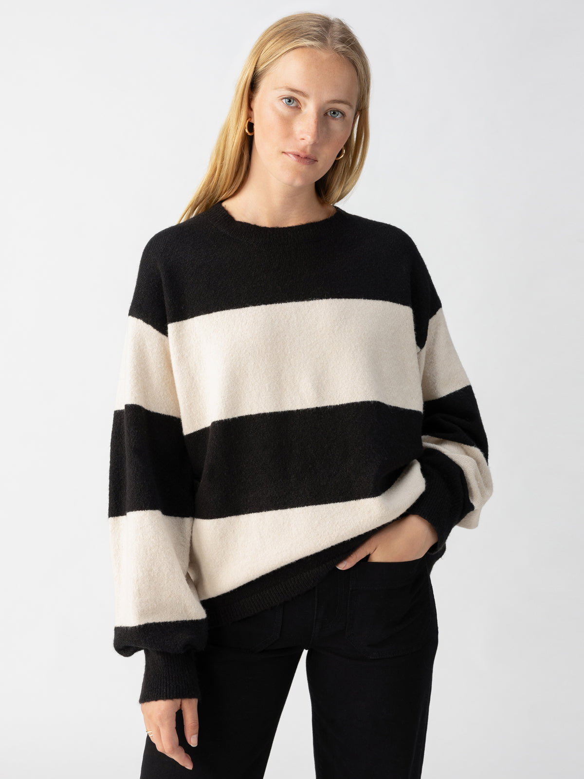 A person stands against a plain white background, wearing the Cuddle Up Crewneck Toasted Almond Black Stripe sweater from Sanctuary Clothing and black pants. The person has long blonde hair, is looking directly at the camera, and has one hand in their pocket.