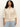 A woman with curly hair wears the Sanctuary Clothing flower pop open stitch jacket in eco natural from their inclusive collection, paired with light blue jeans. She stands against a plain background, looking confidently at the camera.