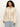 A curly-haired person wears the "flower pop open stitch jacket" from Sanctuary Clothing's eco natural inclusive collection. The cream-colored knit cardigan with floral patterns is open at the collar, over a light shirt and blue jeans. They stand with a neutral expression against a plain background.