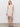 A woman with long blonde hair and a neutral expression stands against a white wall, wearing the Sanctuary Clothing summer shell dress eco natural”a sleeveless, knee-length beige crochet dress”and black sandals.