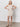 A woman poses against a white wall in the Sanctuary Clothing summer shell dress eco natural, a sleeveless cream crochet, knee-length style, paired with black sandals and her hands on her hips.