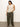 A woman poses against a plain background, wearing Sanctuary Clothing's open stitch shell sweater from the inclusive collection in eco natural over a beige tank. She pairs it with loose-fitting olive-green pants and black sandals, her long dark hair cascading with one hand in her pocket.