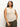 A person with long hair wears a sleeveless open stitch shell sweater in eco natural from the Sanctuary Clothing Inclusive Collection, paired with dark green pants. They are accessorized with a necklace and bracelets, standing against a plain white background, gazing slightly to the side.
