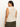 A person with long hair faces away, wearing Sanctuary Clothing's open stitch shell sweater in eco natural from the inclusive collection, paired with loose green pants. They accessorize with gold bracelets on one wrist against a neutral background.