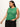 A woman in a Sanctuary Clothing open stitch shell sweater palm inclusive collection, paired with black pants, poses against a plain background. With long brown hair, she accessorizes with hoop earrings and layered necklaces, gazing downwards.