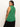 A person with long dark hair turns slightly away, modeling the sleeveless green "Open Stitch Shell Sweater Palm" from Sanctuary Clothing's Inclusive Collection. They're gazing over their shoulder, standing in front of a plain white background and paired with black pants.