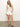 A woman stands against a neutral background in Sanctuary Clothing's open stitch shell sweater eco natural and white shorts, smiling gently with one hand in her pocket. She completes her look with brown strappy sandals.