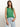 A woman with long brown hair poses against a light backdrop in Sanctuary Clothing's open stitch shell sweater palm and light blue jeans, wearing minimal jewelry. One hand rests in her pocket as she looks into the camera.