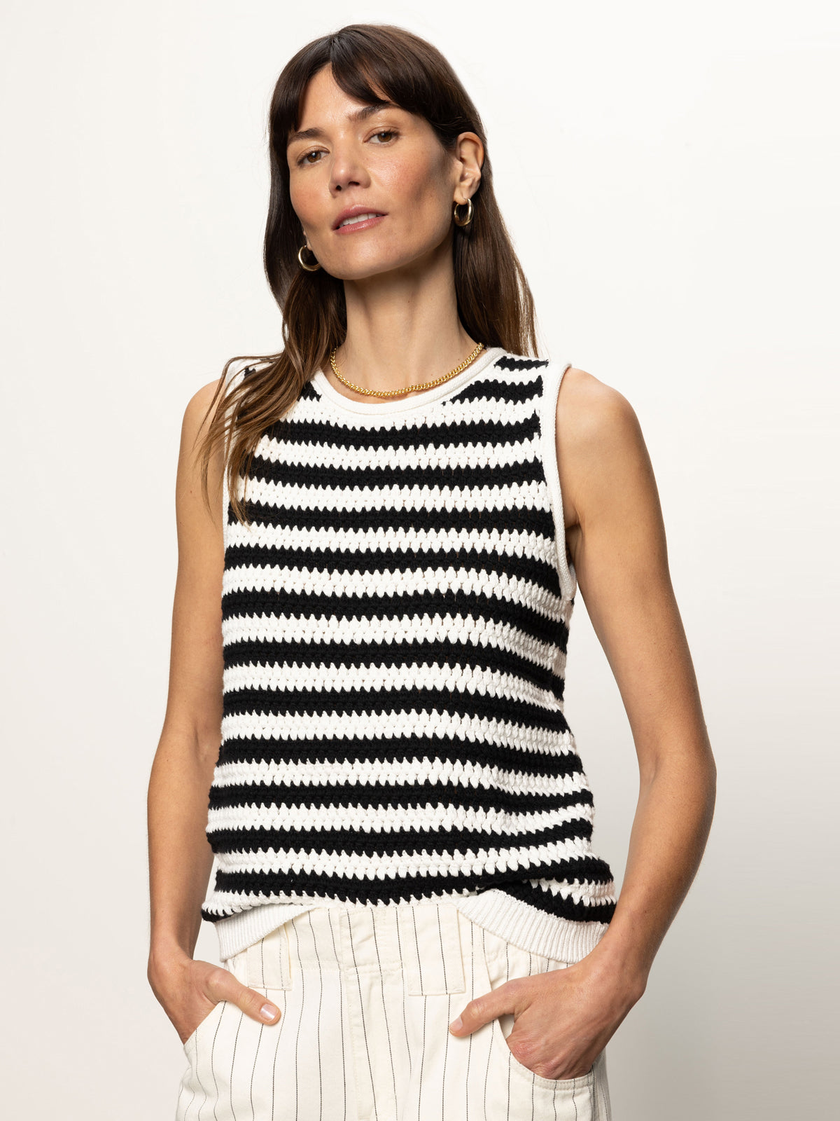 A long-haired woman models a Sanctuary Clothing crochet shell sweater in chalk black stripe, paired with beige striped pants. She poses against a plain background, hands in her pockets, accessorized with hoop earrings and a necklace.