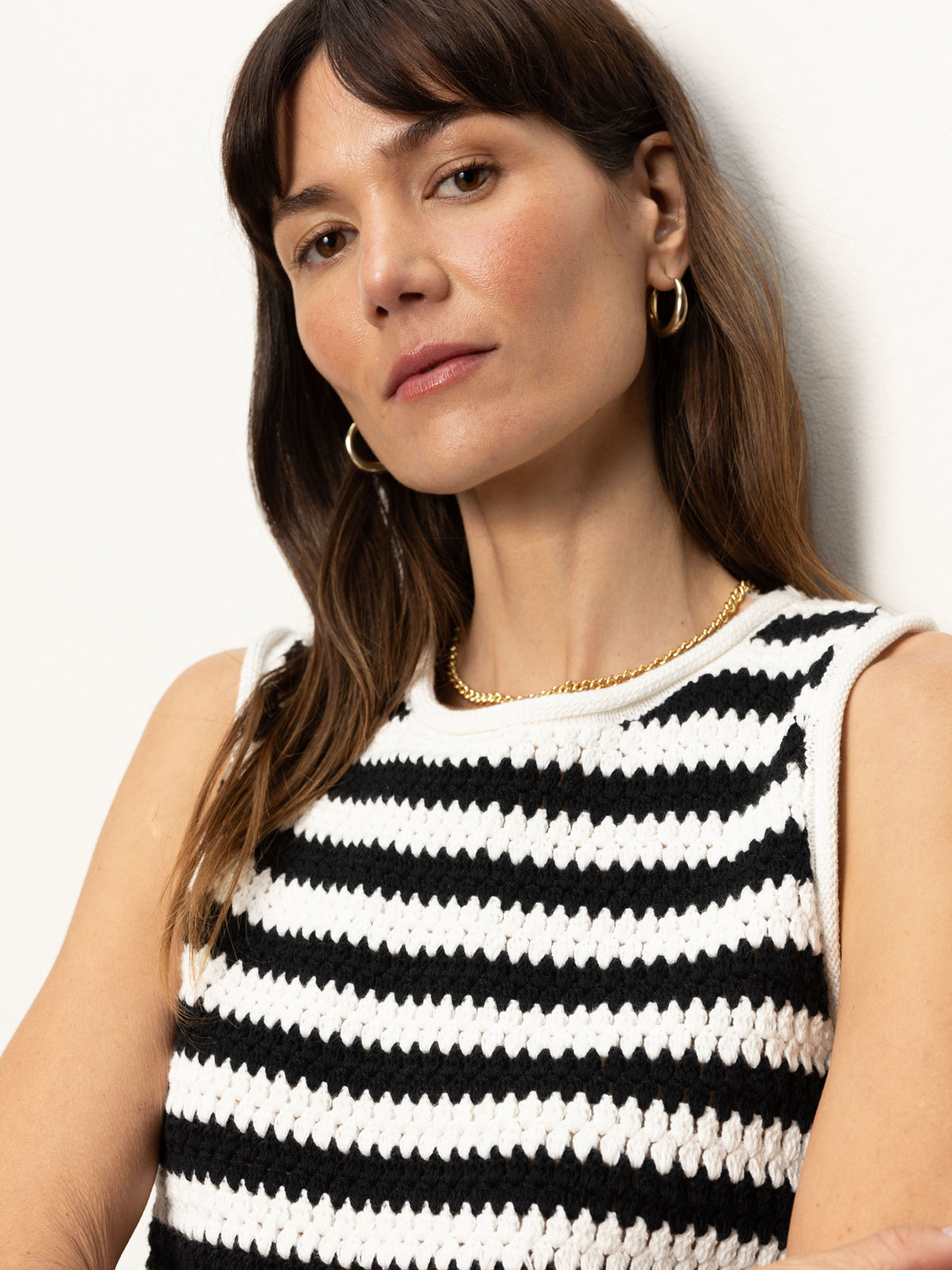 A woman with long brown hair and hoop earrings wears a Sanctuary Clothing crochet shell sweater in chalk black stripe and a gold chain necklace. She stands against a plain white background, looking slightly towards the camera.