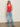 A woman wearing Sanctuary Clothing's mesh stitch shell sweater in cayenne, light blue high-waisted jeans, black sandals, and sunglasses stands against a plain off-white wall with one hand in her pocket.
