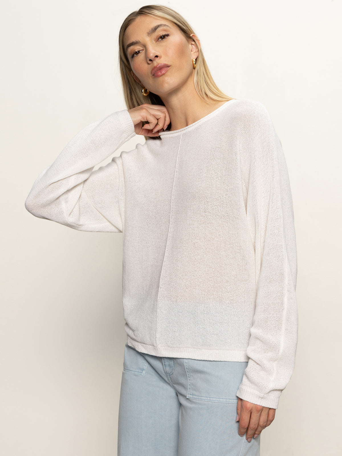 A woman with straight blonde hair models the Sanctuary Clothing easy knitwear top in white and light blue jeans, standing against a plain background with her left hand touching her neck.