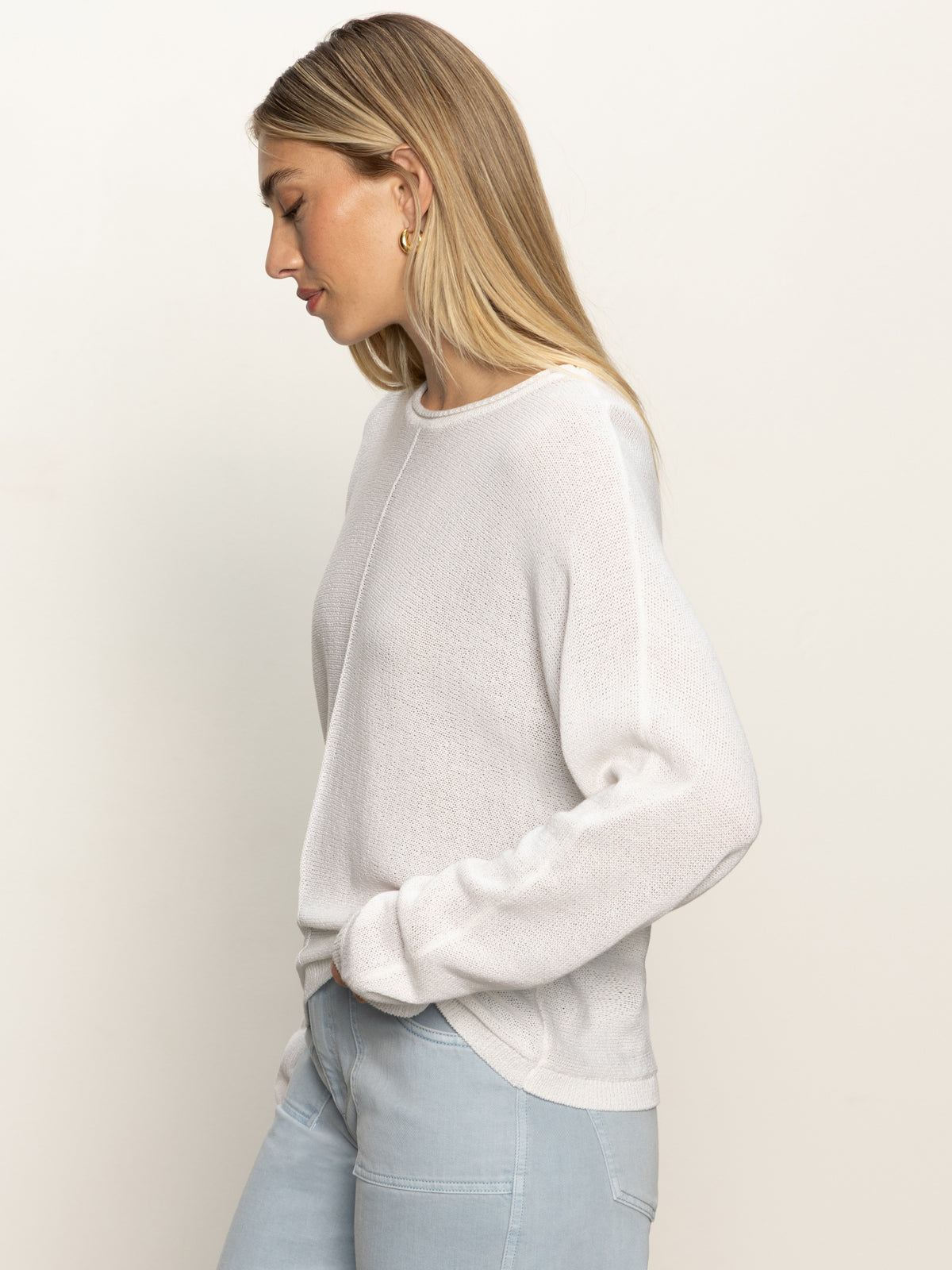 A woman with long blonde hair wears the Sanctuary Clothing easy knitwear top in white and light blue jeans, standing in profile with hands in her pockets against a plain light background.