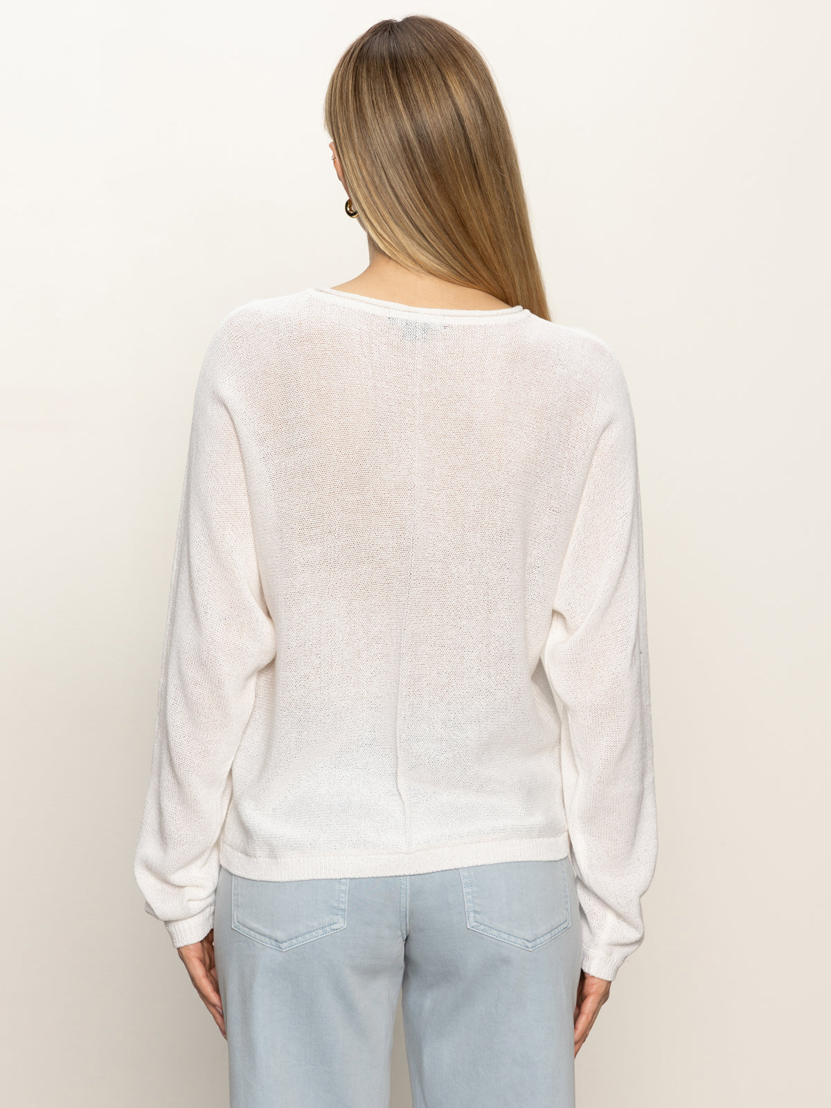 A woman with straight, light brown hair stands with her back to the camera, wearing the Sanctuary Clothing easy knitwear top in white and light blue jeans against a plain off-white background.