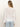 A woman with straight, light brown hair stands with her back to the camera, wearing the Sanctuary Clothing easy knitwear top in white and light blue jeans against a plain off-white background.