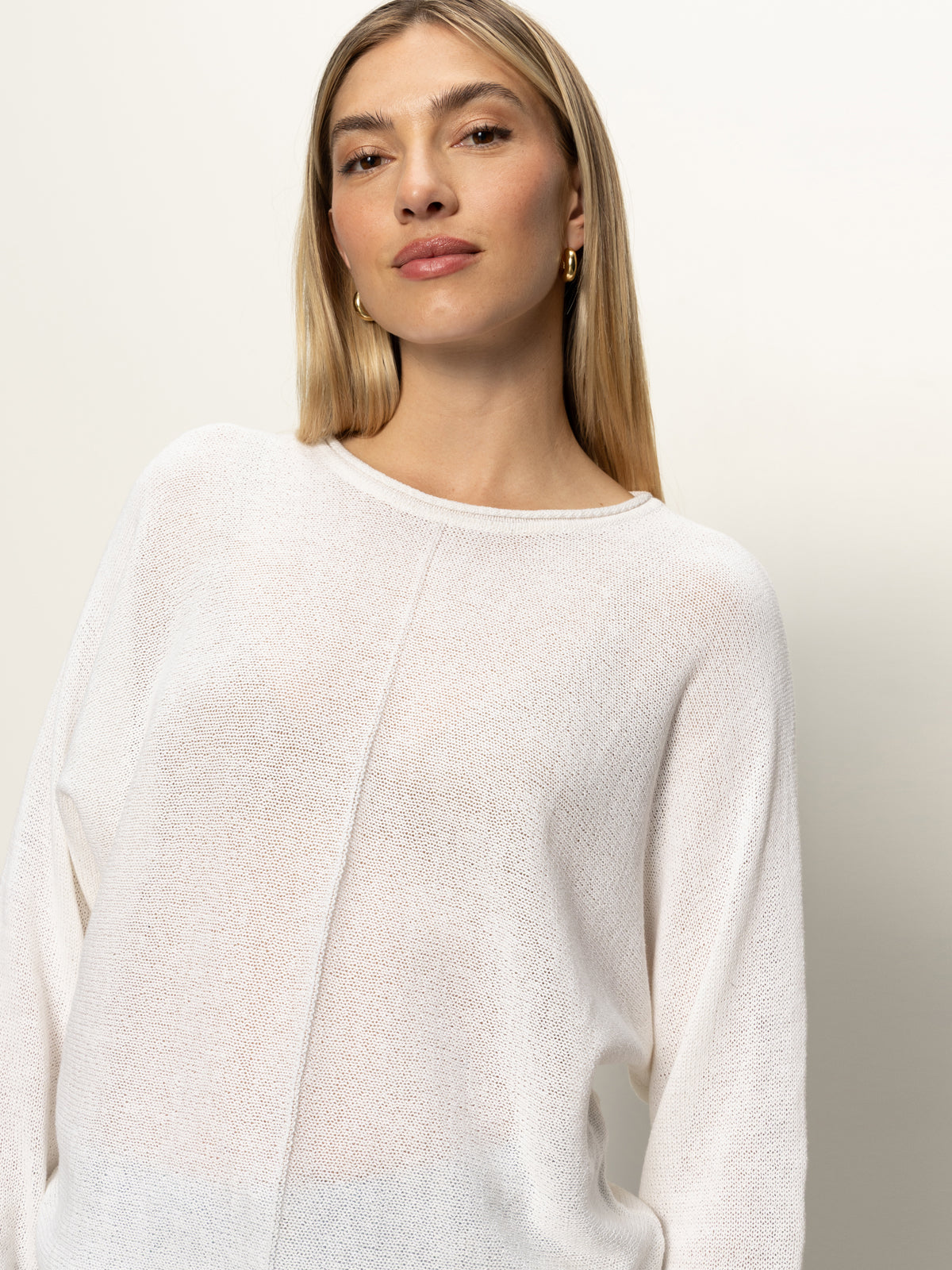 A woman with straight, blonde hair wears the easy knitwear top in white by Sanctuary Clothing and gold hoop earrings, gazing confidently at the camera against a plain light background.