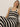 A person in a Sanctuary Clothing crochet sweater skirt in black chalk stripe, sits against a plain wall. With light hair and a neutral expression, they look directly at the camera.