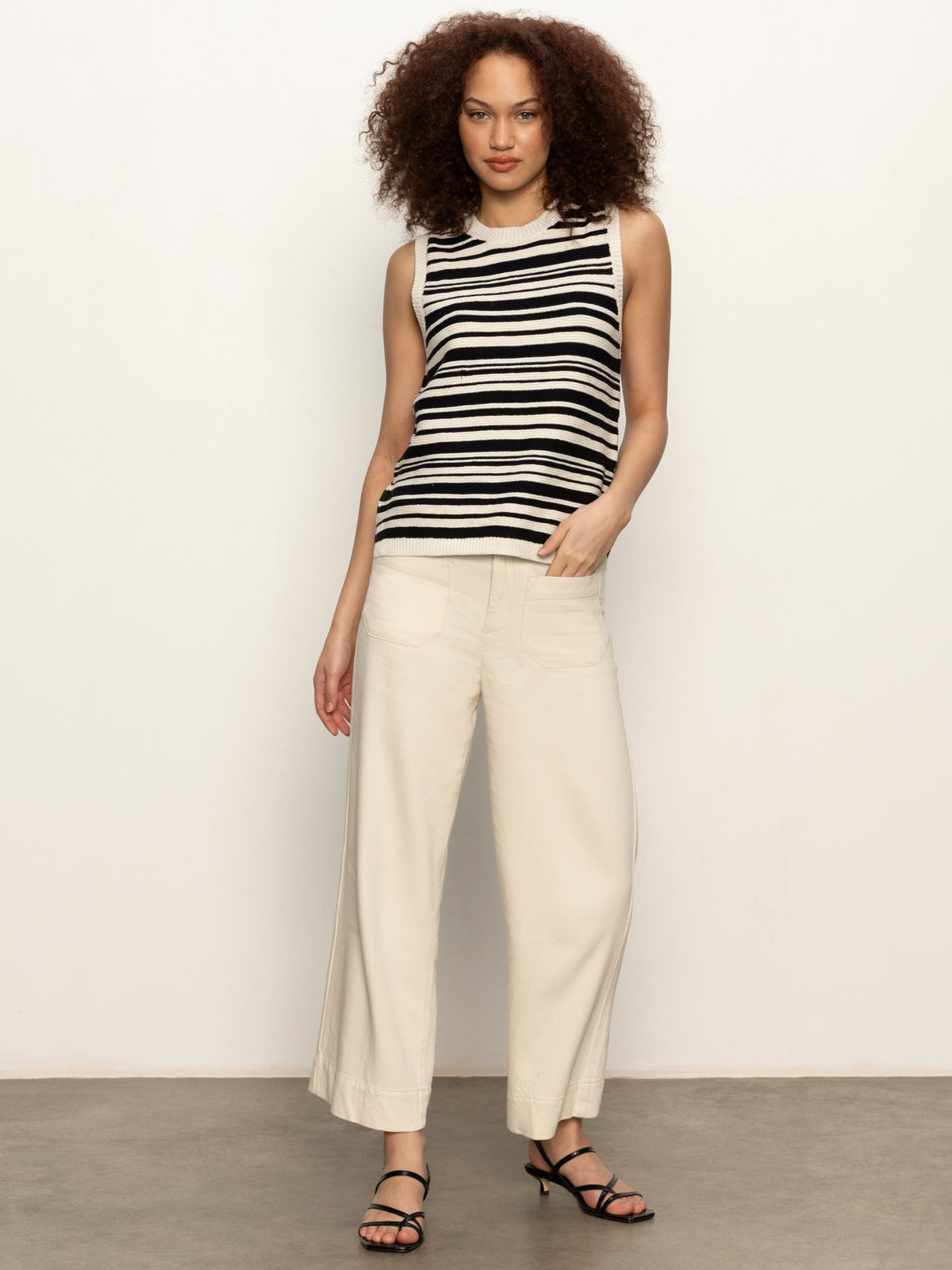A woman with curly hair wears the Sanctuary Clothing modern sweater shell in black macrame, styled with wide-leg cream pants and black strappy sandals, as she stands against a plain white wall.