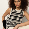 A woman with curly hair sits on a black chair, wearing Sanctuary Clothing’s modern sweater shell in black macrame and light pants, looking confidently at the camera against a plain background.