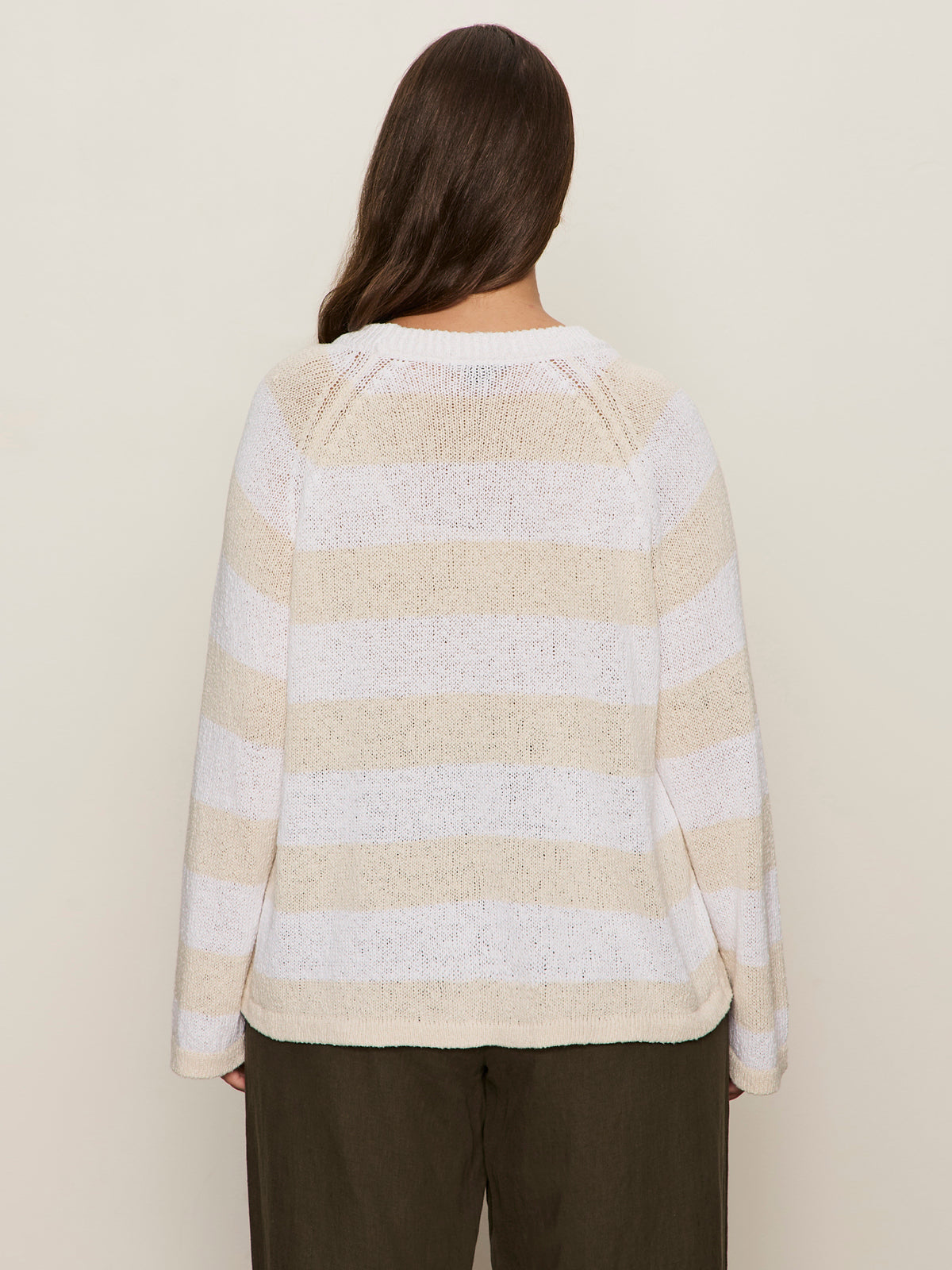 A person with long dark hair is seen from behind wearing the Sanctuary Clothing textured raglan crew white light oat stripe sweater and loose dark pants against a plain light background.
