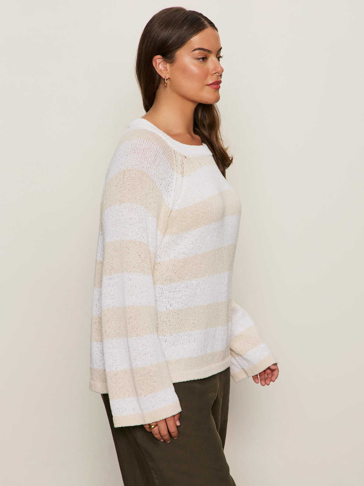 A woman with long, wavy brown hair wears Sanctuary Clothing’s textured raglan crew in white light oat stripe (extended sizing) and dark olive pants, accessorized with small hoop earrings and rings, posed in profile against a plain background.