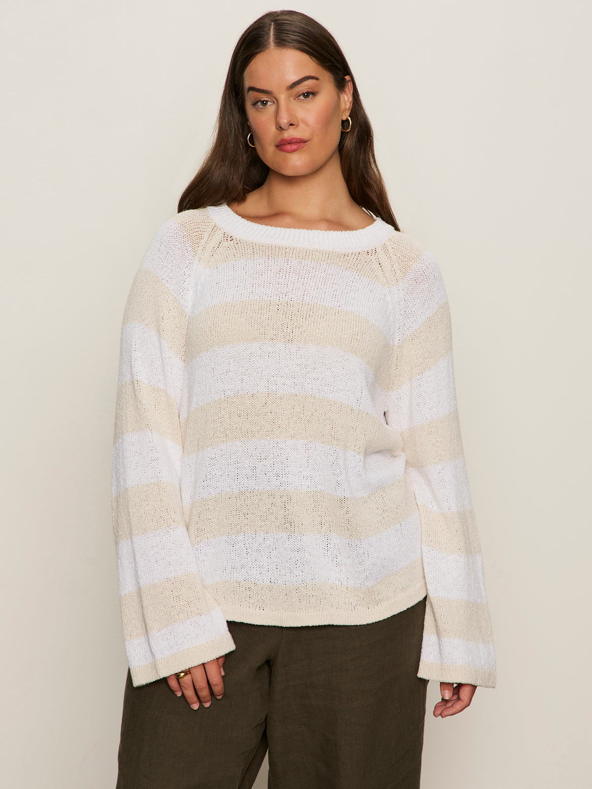 A woman with long brown hair models the Sanctuary Clothing textured raglan crew in white light oat stripe (extended sizing), featuring wide sleeves. She pairs it with dark pants and stands against a plain, light background, looking confidently at the camera.