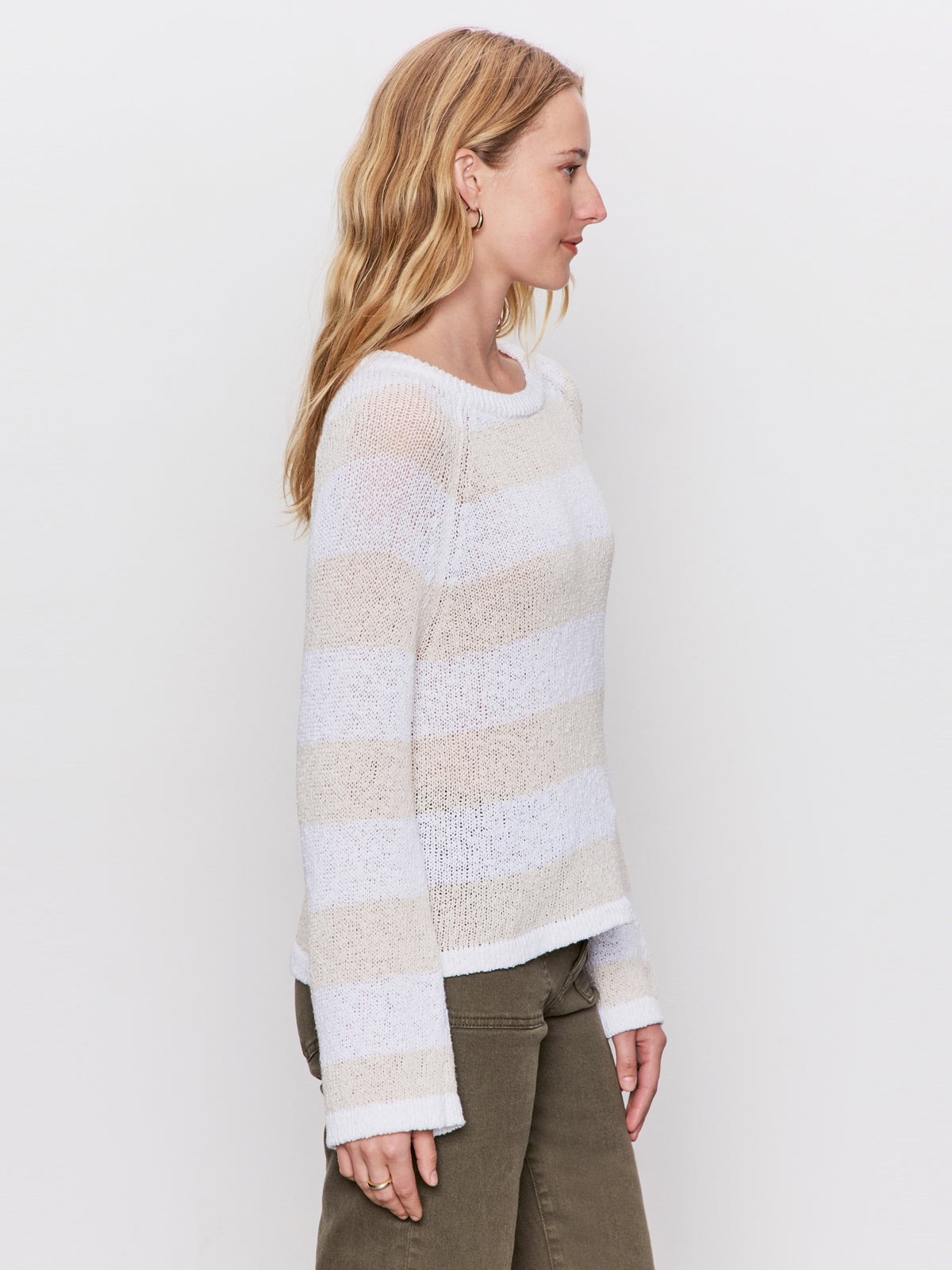 A woman with long blonde hair stands in profile against a white background, wearing Sanctuary Clothing’s textured raglan crew in white light oat stripe and olive green pants.