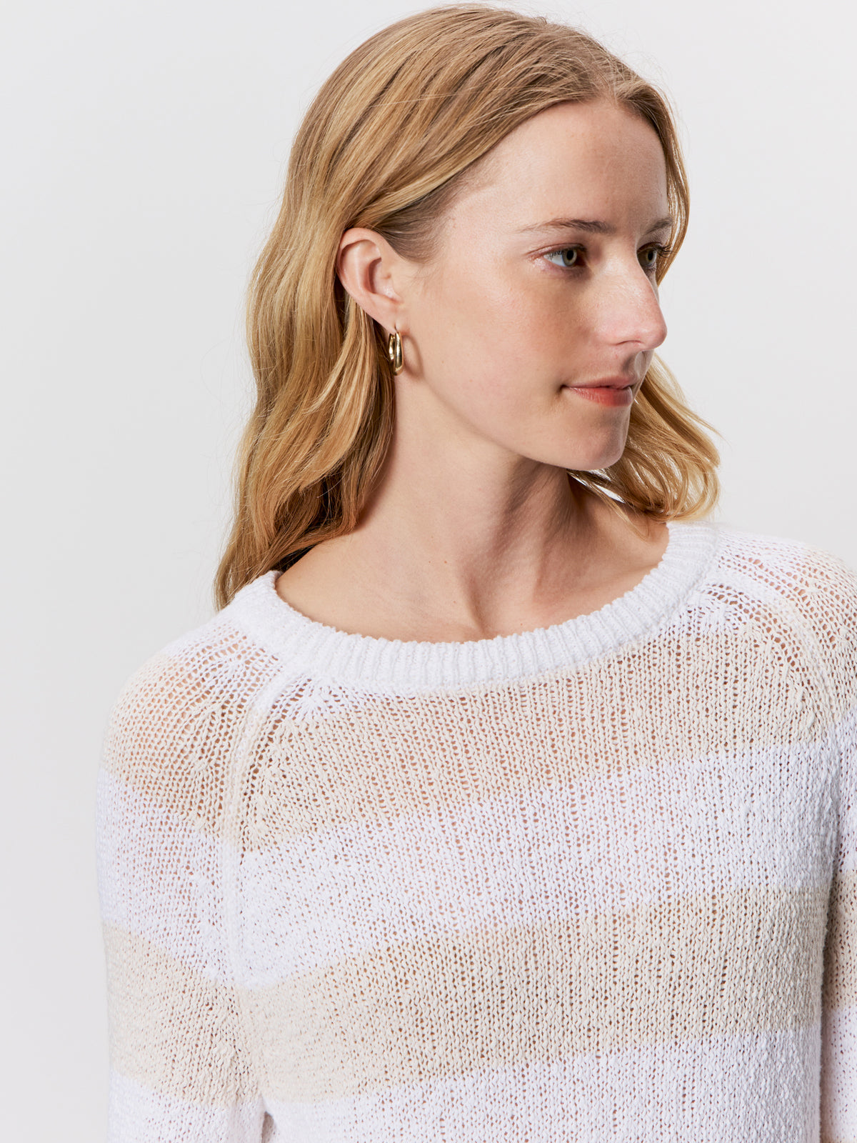 A woman with wavy blonde hair and gold hoop earrings models the Sanctuary Clothing textured raglan crew in white light oat stripe, looking to the side against a plain light background.