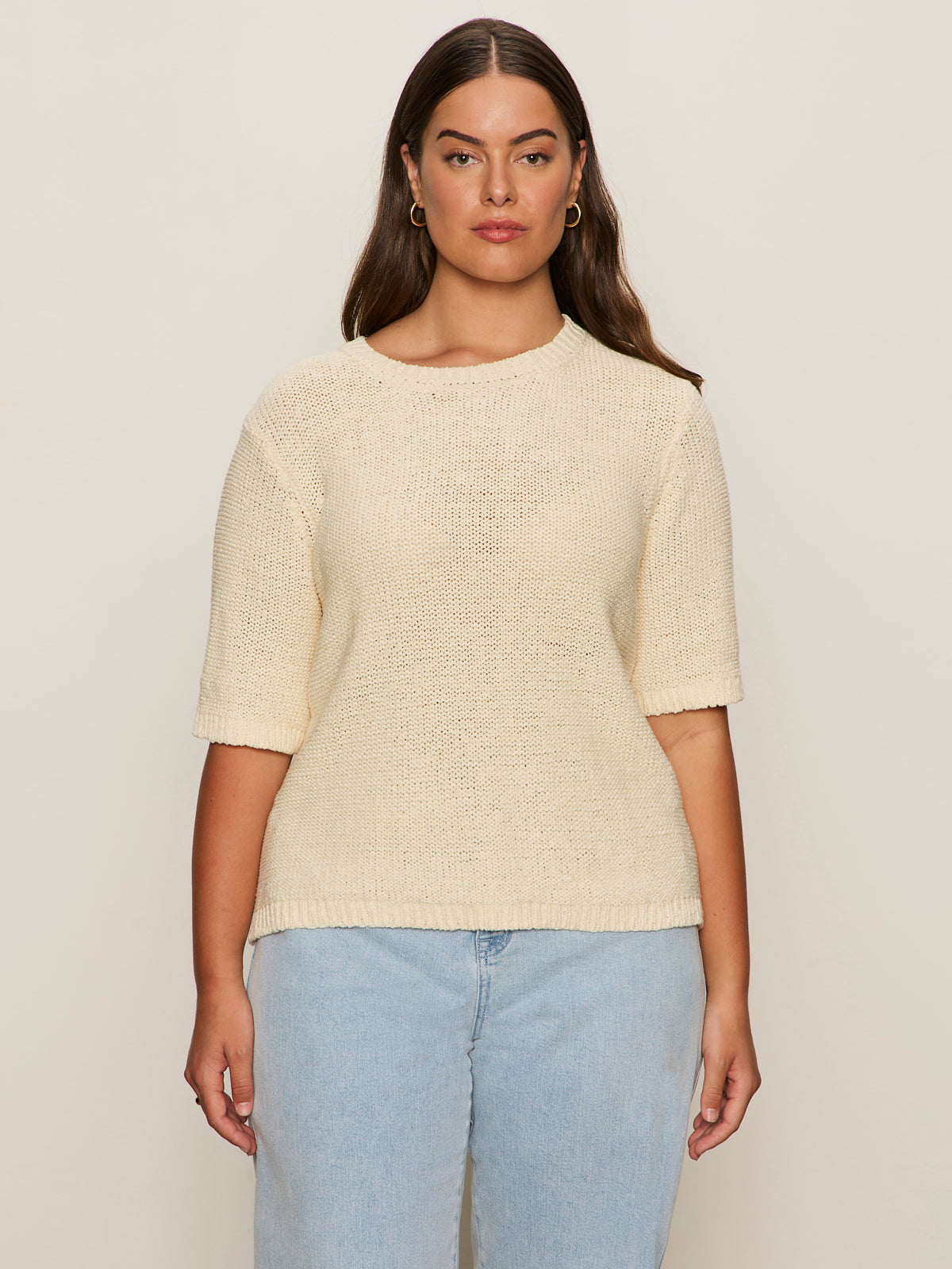 A woman with long brown hair wears the Sanctuary Clothing tape yarn tee macrame (extended sizing), a cream knit short-sleeve sweater, paired with light blue jeans, standing against a plain beige background.