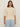 A woman with long brown hair wears the Sanctuary Clothing tape yarn tee macrame (extended sizing), a cream knit short-sleeve sweater, paired with light blue jeans, standing against a plain beige background.
