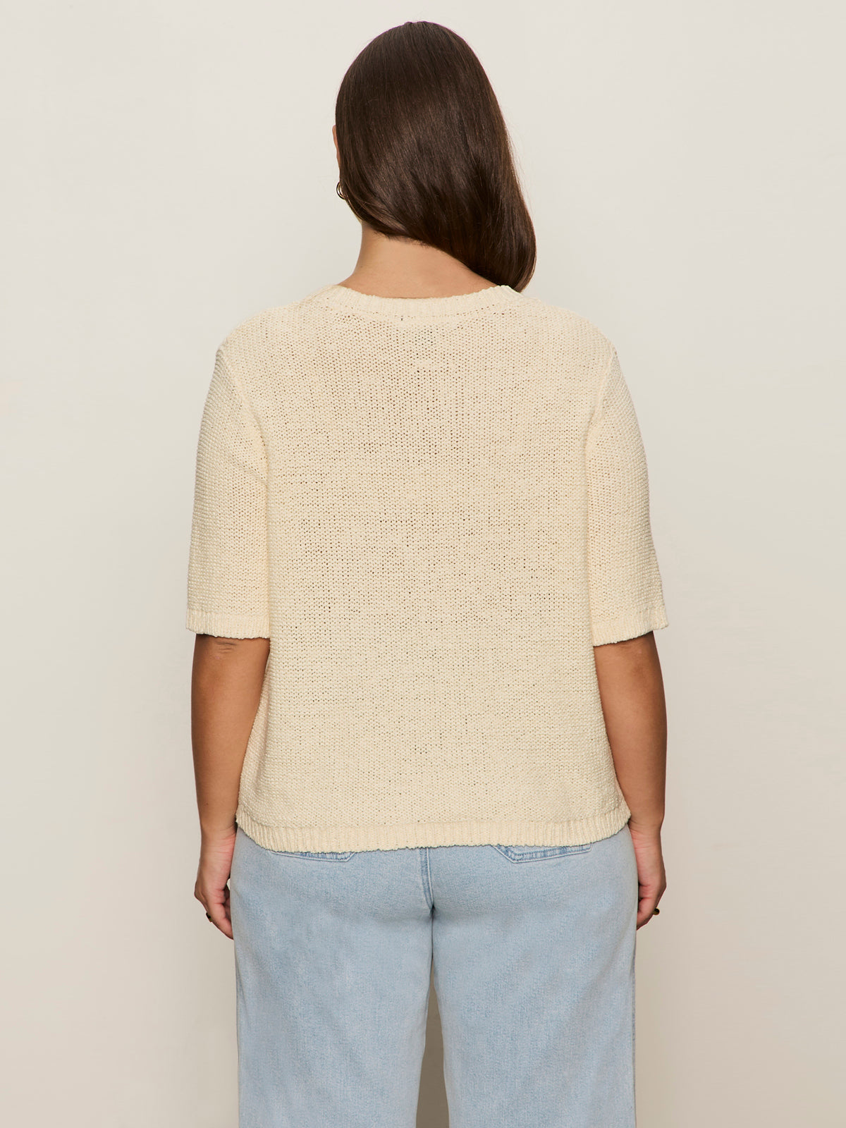 A person with long brown hair wears the Sanctuary Clothing tape yarn tee macrame in cream and light blue jeans, shown from the back against a beige background.