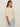 A woman with long brown hair stands in profile against a plain light background, wearing Sanctuary Clothing's tape yarn tee macrame extended sizing and light blue jeans.