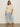 A woman with long brown hair poses confidently in a Sanctuary Clothing tape yarn tee macrame (extended sizing), paired with light blue wide-leg cropped jeans and cream slide sandals, standing against a plain wall.