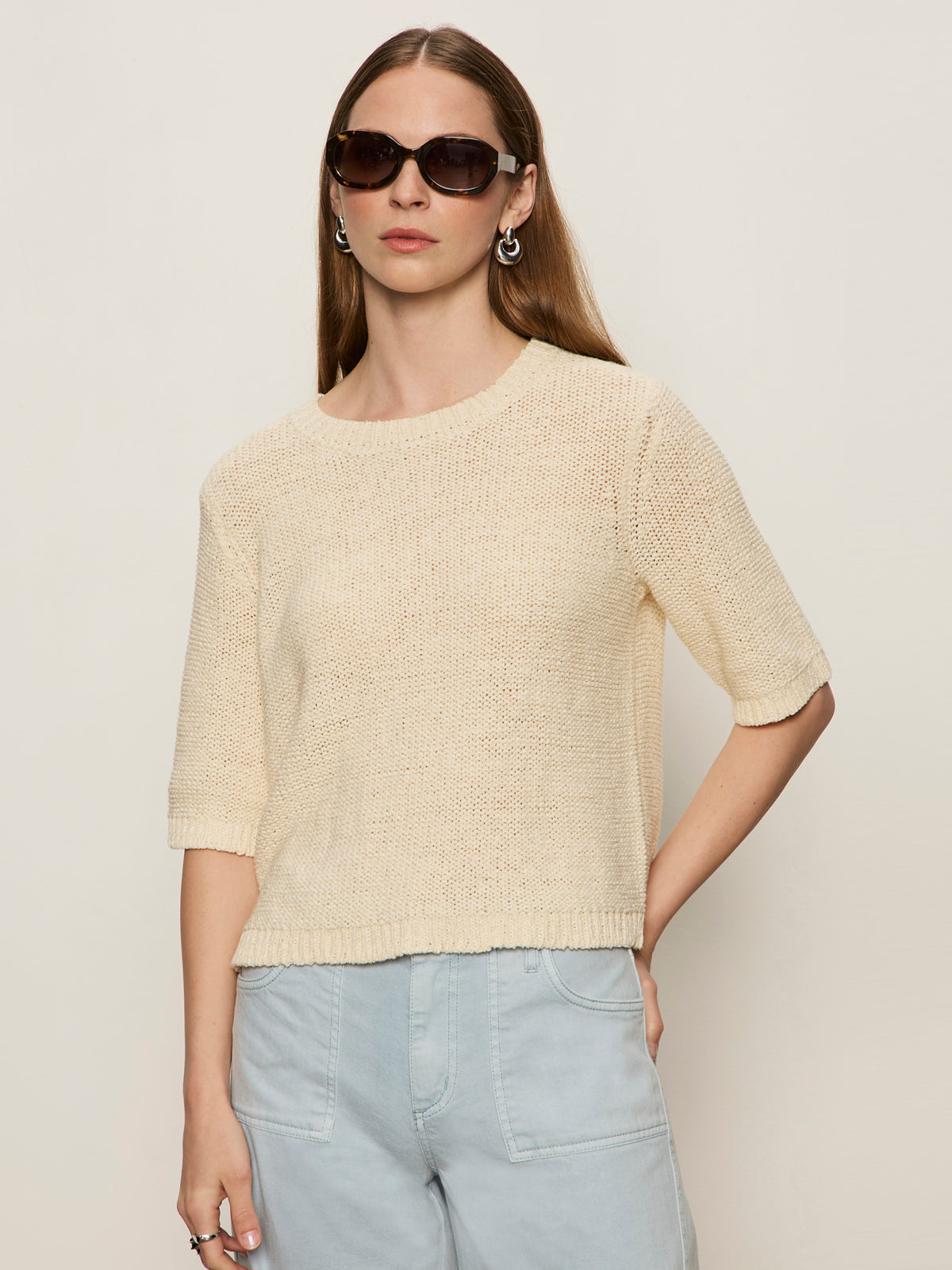 A woman with long brown hair wears dark sunglasses, silver hoop earrings, and the Sanctuary Clothing tape yarn tee macrame—a cream short-sleeve knit sweater—paired with light blue high-waisted pants against a plain light background.