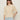 A woman with long brown hair wears dark sunglasses, silver hoop earrings, and the Sanctuary Clothing tape yarn tee macrame—a cream short-sleeve knit sweater—paired with light blue high-waisted pants against a plain light background.