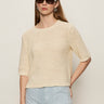 A woman with long brown hair wears dark sunglasses, silver hoop earrings, and the Sanctuary Clothing tape yarn tee macrame—a cream short-sleeve knit sweater—paired with light blue high-waisted pants against a plain light background.