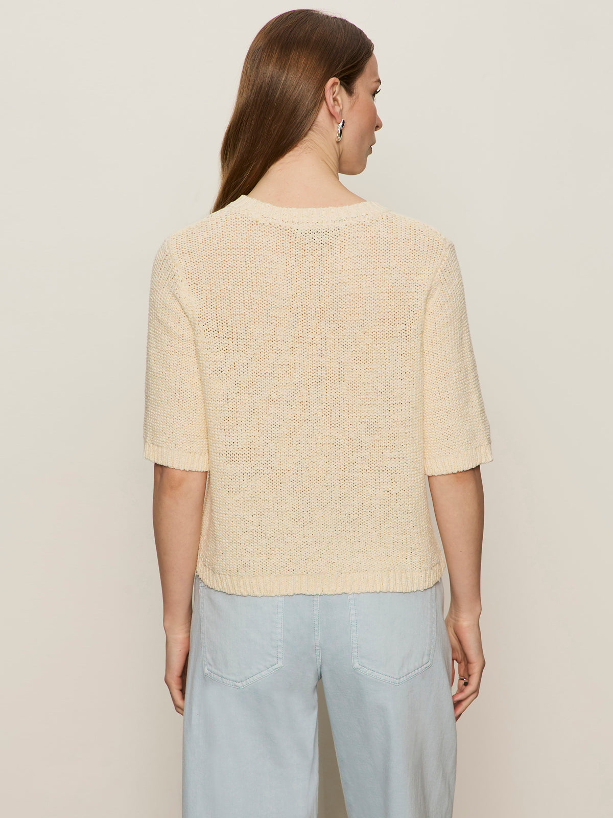 A woman with long brown hair, seen from the back, wears the Sanctuary Clothing tape yarn tee macrame and light blue high-waisted pants against a beige background.