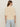 A woman with long brown hair, seen from the back, wears the Sanctuary Clothing tape yarn tee macrame and light blue high-waisted pants against a beige background.