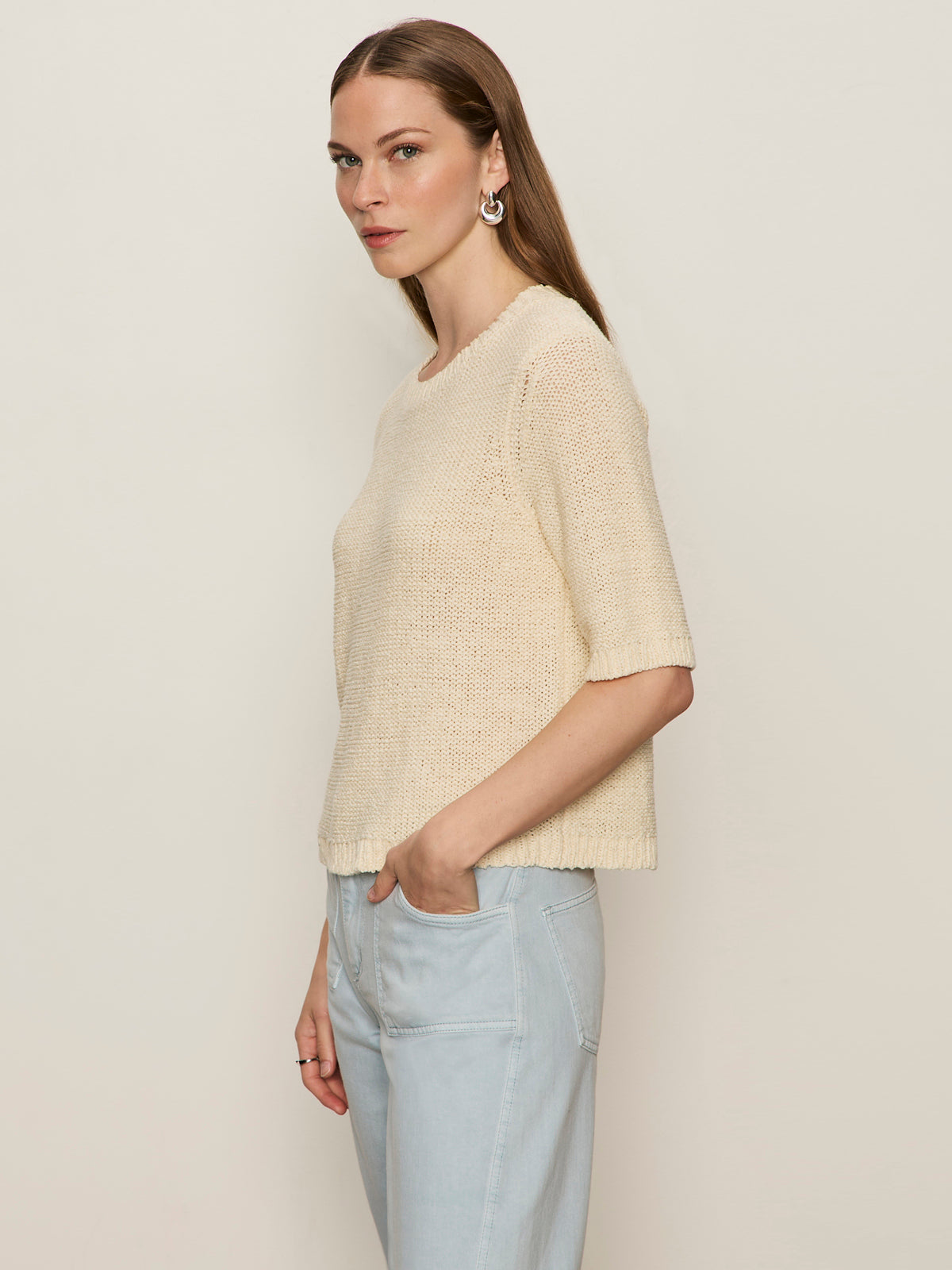 A woman with long brown hair stands sideways against a plain light background, wearing Sanctuary Clothing's tape yarn tee macrame in cream and light blue high-waisted jeans, with one hand in her pocket.