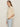 A woman with long brown hair stands sideways against a plain light background, wearing Sanctuary Clothing's tape yarn tee macrame in cream and light blue high-waisted jeans, with one hand in her pocket.