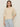 A woman with straight brown hair wears silver hoop earrings and the Sanctuary Clothing tape yarn tee macrame, a cream short-sleeve knit sweater, paired with light blue high-waisted jeans against a beige background.