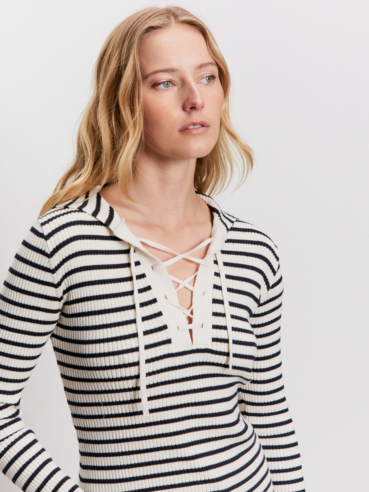A woman with long blonde hair wears a cream and black striped ribbed top with a lace-up V-neck. She looks slightly to the side against a plain white background.
