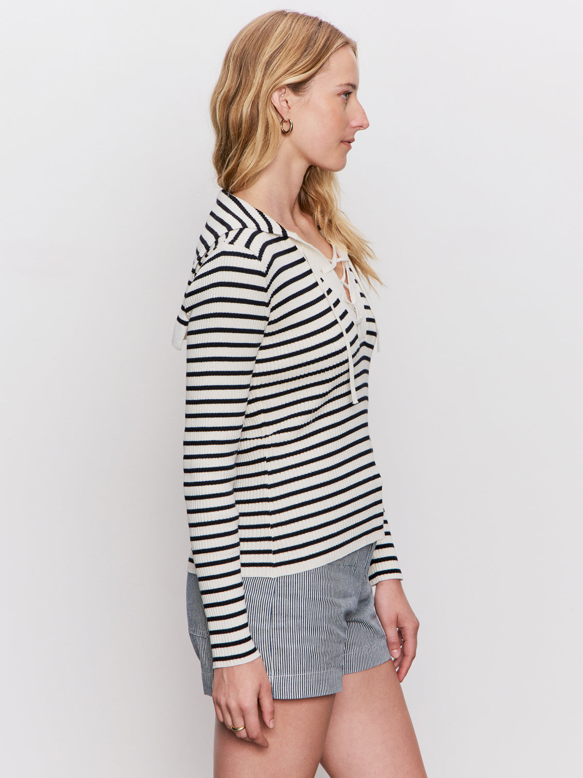 A woman with blonde hair stands in profile wearing a black-and-white striped long-sleeve top with a lace-up neckline and gray pinstriped shorts against a plain white background.