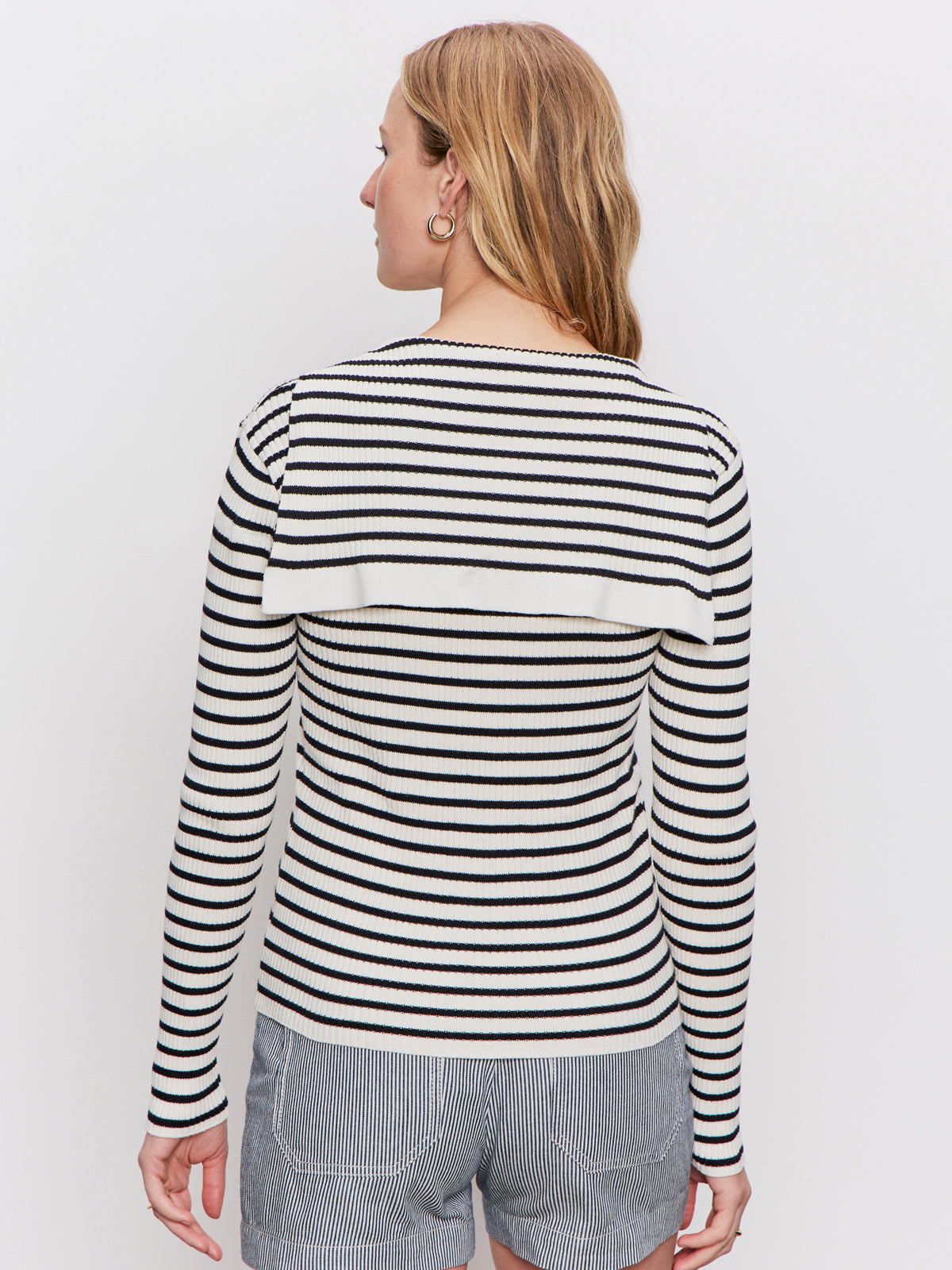 A woman with light brown hair stands facing away, wearing a long-sleeve, black-and-white striped top with a flap detail on the back and gray-and-white striped shorts against a plain white background.