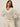 A woman with long brown hair wears the Sanctuary Clothing airy openwork sweater macrame in cream, paired with cream belted pants. She stands against a plain light background, hands on her hips, looking confidently at the camera.