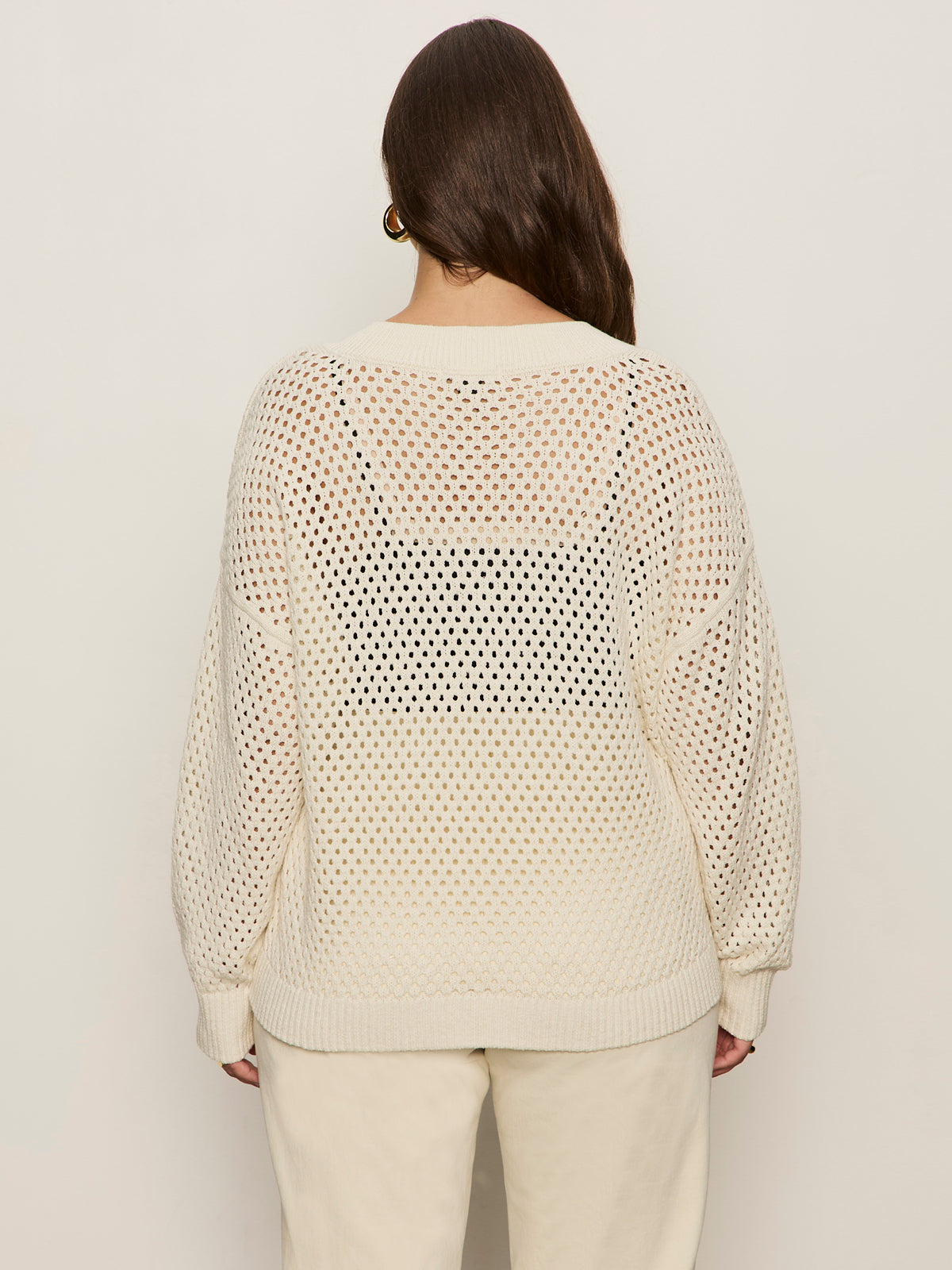 A person with long brown hair is seen from behind wearing the Sanctuary Clothing airy openwork sweater macrame (extended sizing) in cream over a black top and matching cream pants against a plain background.