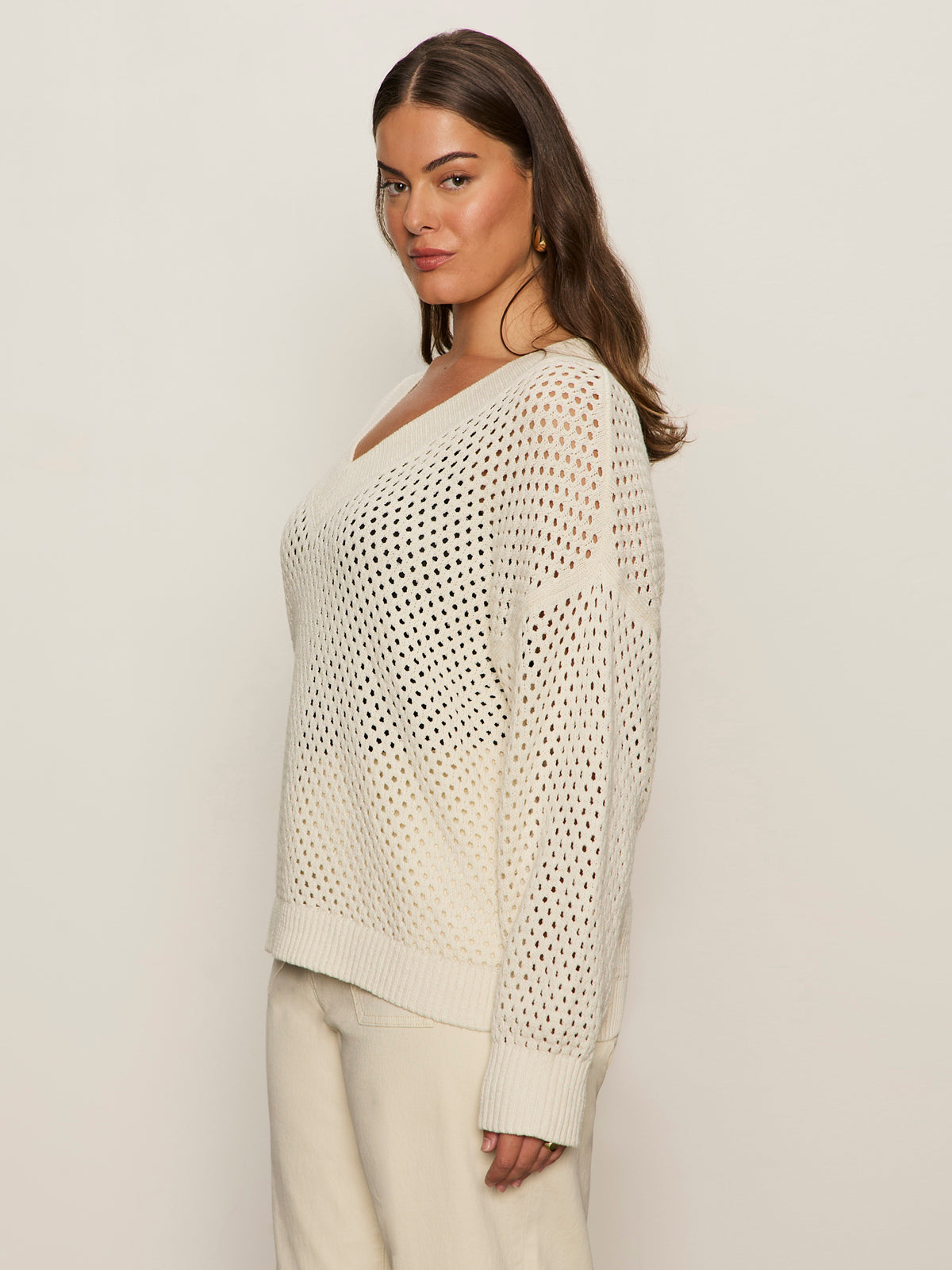 A woman with long brown hair models the Sanctuary Clothing airy openwork sweater macrame in cream, paired with light pants. She stands sideways, looking at the camera against a plain, light background. Extended sizing available.