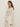 A woman with long brown hair models the Sanctuary Clothing airy openwork sweater macrame in cream, paired with light pants. She stands sideways, looking at the camera against a plain, light background. Extended sizing available.