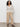 A woman with curly hair stands against a plain background, hands in pockets, wearing the Sanctuary Clothing airy openwork sweater macrame, beige wide-leg pants, and black strappy sandals.
