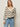 A woman with straight blonde hair wears a cream sweater with black horizontal stripes and light blue jeans. She has one hand in her pocket and wears hoop earrings, standing against a plain light background.